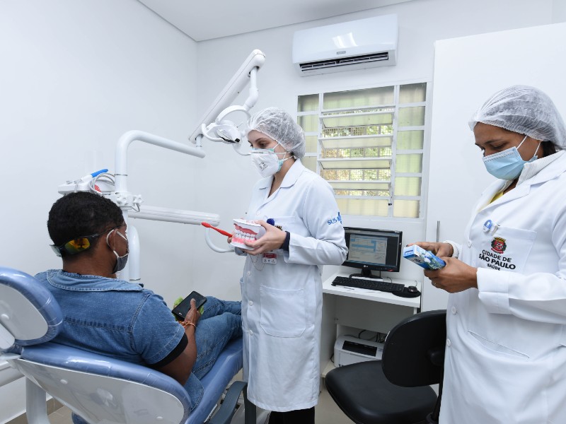 Um paciente com camisa jeans está sentado na cadeira de dentista. Em pé na frente dele, a dentista segura um molde de uma boca e uma escova de dentes. Atrás dela está a assistente. Ambas estão de jaleco branco, toca e máscara